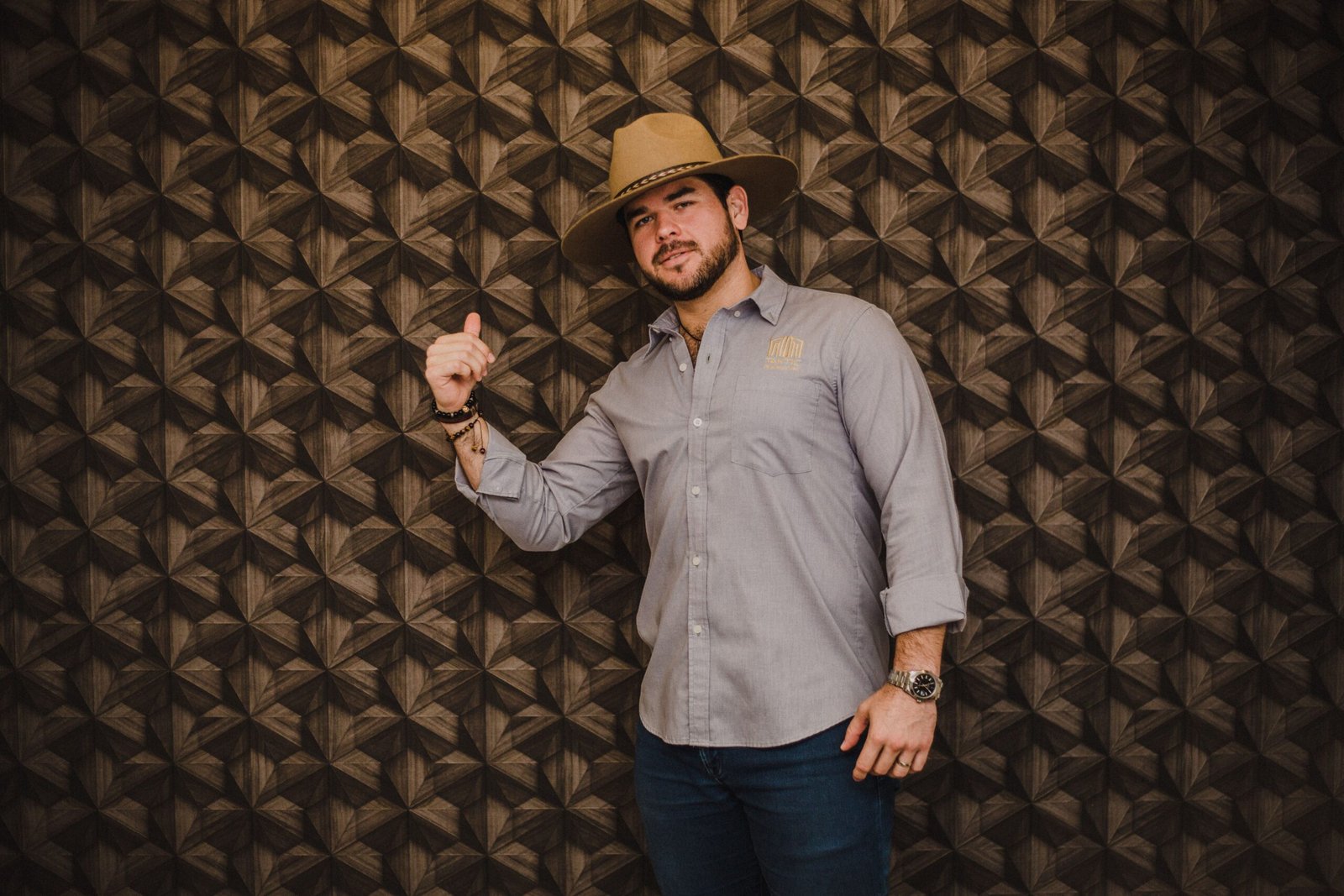 Confident man in a hat posing against a patterned wall indoors.